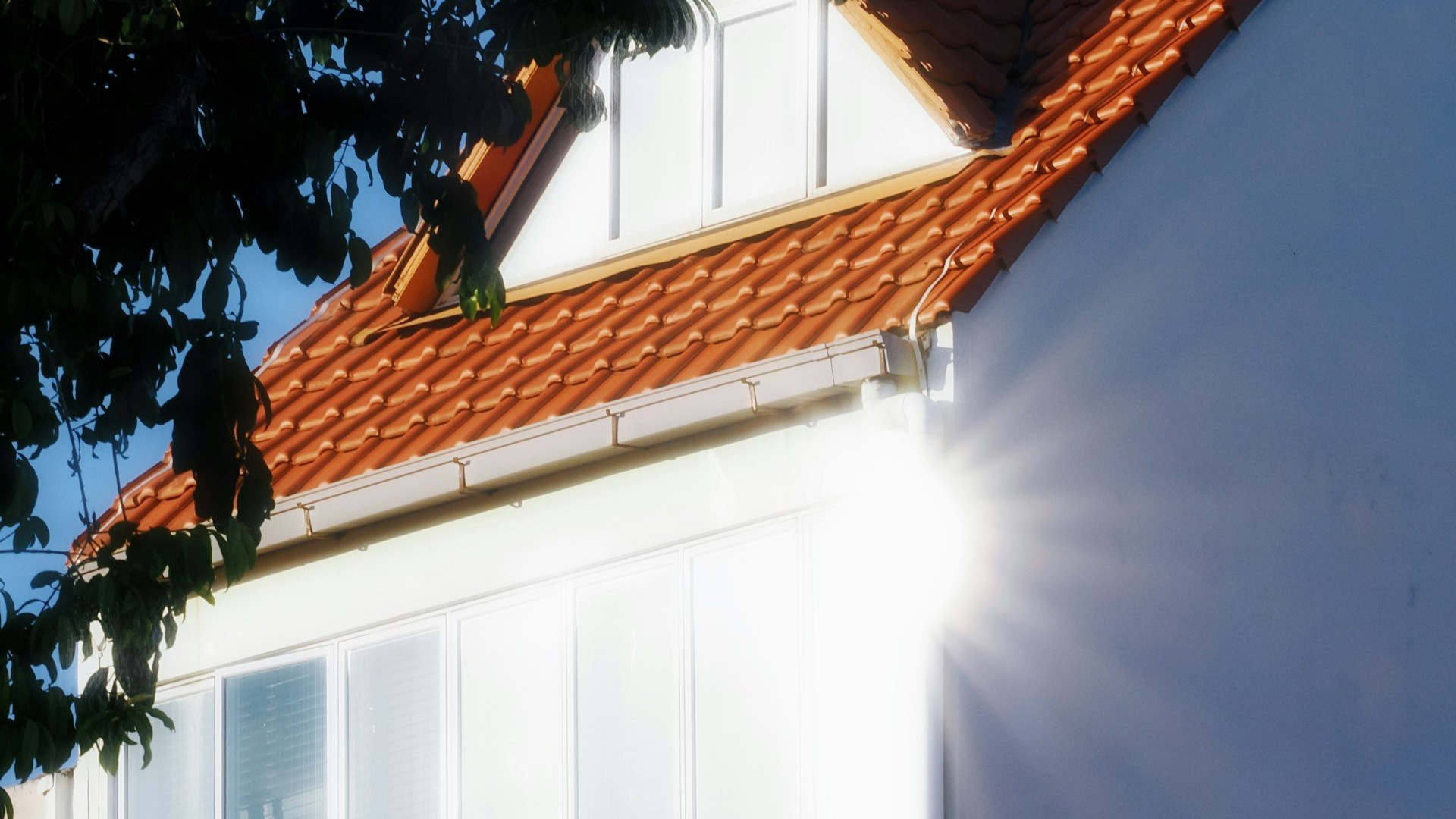 Fel zonlicht valt op de grote glazen pui van een woning met rood pannendak, waardoor de ramen sterk oplichten